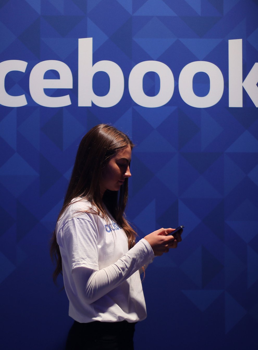 A woman looks at her smartphone in front of a Facebook logo