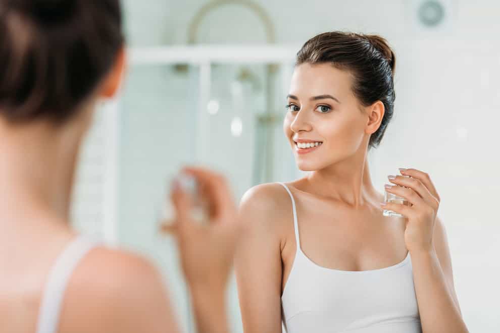 beautiful young woman holding perfume bottle and looking at mirror in bathroom