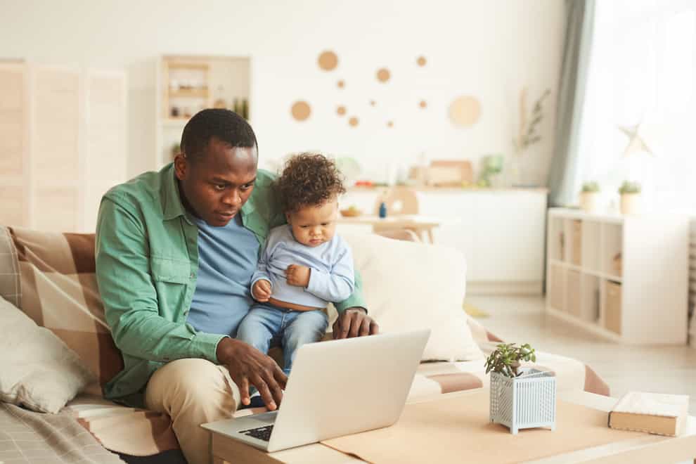 A middle-aged black man sitting on the sofa at home with a toddler son on his lap, looking at a laptop