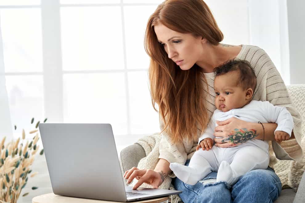 Busy mother using laptop working from home with little african baby daughter.