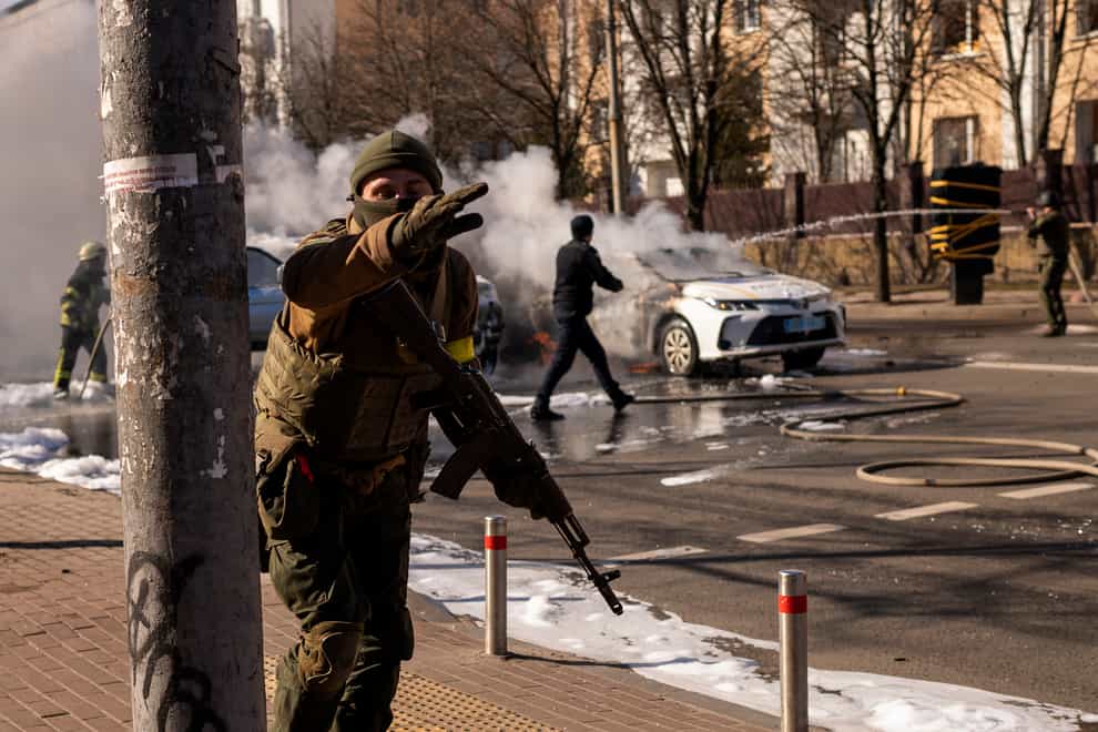 Ukrainian soldiers take positions outside a military facility as two cars burn, in a street in Kyiv (AP)