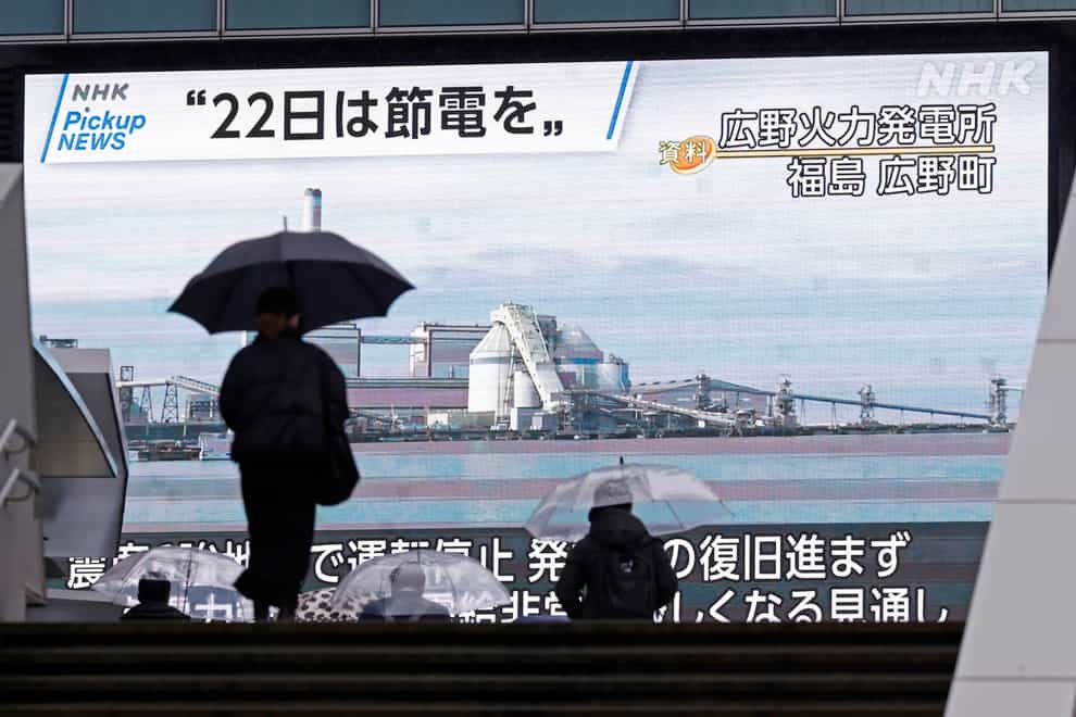 A woman with an umbrella walks past a monitor calling for people to conserve electricity (Kyodo News via AP)