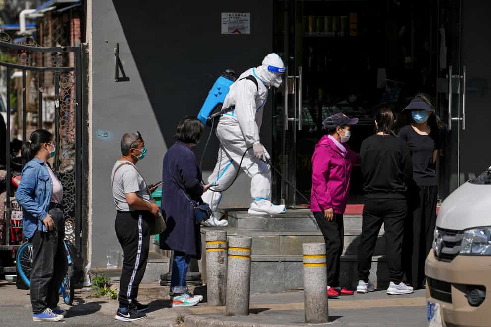 A worker in a protective suit sprays disinfectant as residents wearing face masks line up for mass coronavirus testing outside a residential complex on Tuesday May 3 2022 in Beijing (Andy Wong/AP)