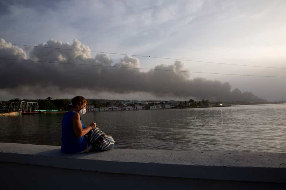 A resident sits on the sea wall as smoke rises in the background from a deadly fire at a large oil storage facility in Matanzas, Cuba (Ismael Francisco/AP)