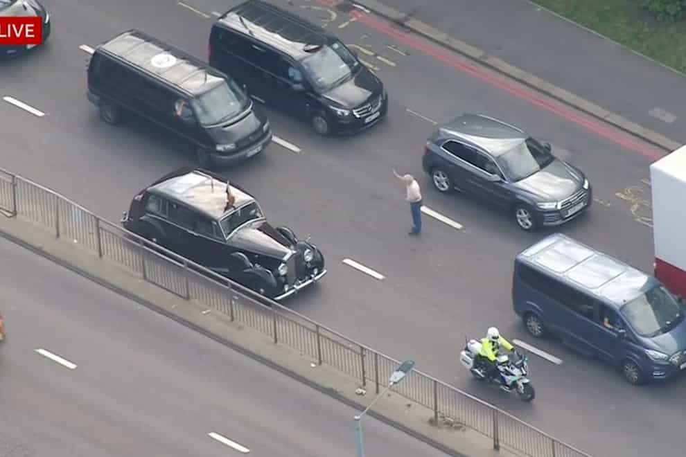 Screengrab from BBC News of a member of the public stepping out into the road towards a car carrying King Charles III and the Queen Consort to take a photo with close protection officers leaving a car which is providing security and gesturing to the member of public (BBC/PA)