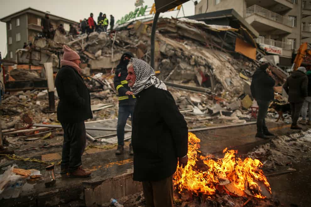 People warm themselves next to a collapsed building in Malatya earlier this month (AP)