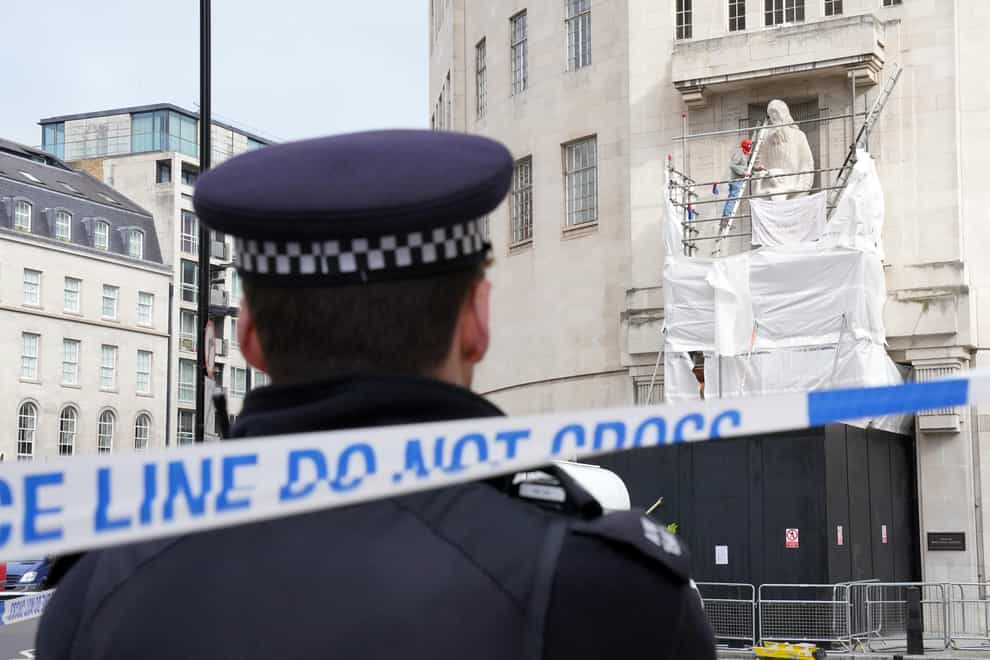 A police officer watches as a man wearing a Spiderman mask uses a hammer and chisel to damage the Prospero and Ariel statue by Eric Gill, outside BBC Broadcasting House in London, in an apparent protest. Picture date: Saturday May 20, 2023.